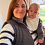 Daniel is registered to the contest to win money with this photo: woman, baby, indoor, smile, holding, bib, window, natural_light, clothing, curious, striped_sweater, vest, person, portrait, family, casual, face, head, home, daylight