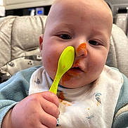 Daniel joined the competition — help win amazing prizes! baby, child, spoon, messy_face, bib, high_chair, food, orange_food, hand, clothing, indoor, kitchen, person, face, cushion, feeding, closeup, cute, expression, toddler
