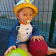 Liam a rejoint le concours — aidez-le/la à gagner de superbes lots ! toddler, child, hat, pacifier, ball, green_ball, red_ball, sitting, outdoor, fence, clothing, shorts, long_sleeve_shirt, barefoot, play, toy, baby, young_child, person, daylight