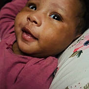 Milani is registered to the contest to win money with this photo: baby, infant, face, smiling, eyes, pink_clothing, pillow, floral_pattern, closeup, cute, child, portrait, skin, lying_down, indoors, young, head, expression, person, adorable