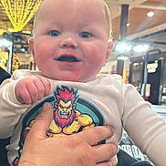 Ethan is registered to the contest to win money with this photo: baby, infant, child, person, hand, clothing, cartoon_print, indoor, lights, smiling, cute, holding, sitting, portrait, face, adult_hand, watch, long_sleeve, happy, casual