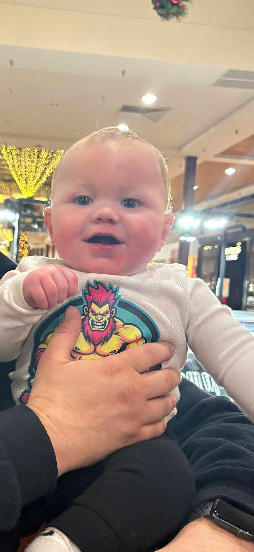 Ethan is registered to the contest to win money with this photo: baby, infant, child, person, hand, clothing, cartoon_print, indoor, lights, smiling, cute, holding, sitting, portrait, face, adult_hand, watch, long_sleeve, happy, casual