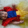 apple, baby, blue_bodice, child, close_up, costume, fairytale, family_photography, fluffy_rug, gold_decoration, happy, indoor, infant, lying_down, portrait, princess_costume, red_bow, smiling, white_background, yellow_dress