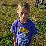 Bentley is registered to the contest to win money with this photo: child, boy, field_trip, purple_shirt, outdoor, grass, daylight, group, kids, casual_clothing, standing, serious_expression, elementary_school, name_tag, short_hair, summer, people, children, school_event, sunlight