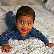 Kyra is registered to the contest to win money with this photo: baby, child, infant, smiling, lying_down, blue_clothing, polka_dot, bed, bedspread, floral_pattern, hands, face, eyes, happy, cute, indoor, portrait, person, young_child, skin