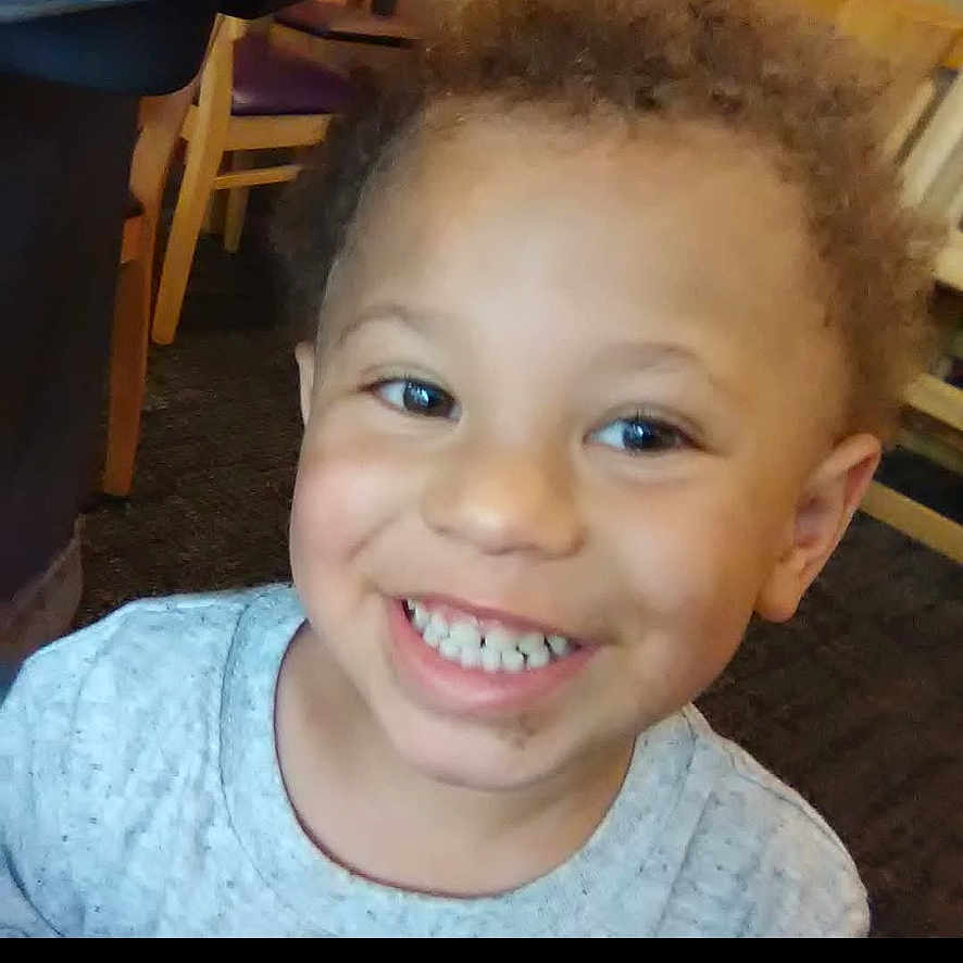 Le'Andre is registered to the contest to win money with this photo: background, casual_clothing, child, curly_hair, cute, expression, face, happy, headshot, indoor, joyful, light_skin, person, portrait, sitting, smiling, teeth, toddler, wooden_chair, young_child