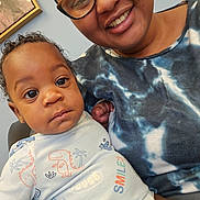 Lamontè joined the competition — help win amazing prizes! adult, baby, chair, closeup, clothing, curly_hair, dinosaur, face, frame, glasses, happy, indoor, onesie, person, portrait, seated, skin, smile, tie_dye, wall_art