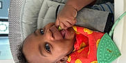 Lamontè is registered to the contest to win money with this photo: baby, baby_food, bib, blue_shirt, child, closeup, curly_hair, cute, face, feeding, hand, high_chair, indoor, infant, orange_bib, person, playful, smiling, table, teething_toy