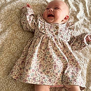 Victoire a rejoint le concours — aidez-le/la à gagner de superbes lots ! baby, child, smiling, floral_dress, white_socks, white_shoes, patterned_blanket, lying_down, happy, infant, cute, portrait, indoors, soft_lighting, human, young_child, clothing, baby_leg, baby_face, baby_hand