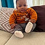 baby, child, smiling, orange_sweater, gray_pants, socks, couch, pillows, patterned_pillow, solid_pillow, indoor, curtains, natural_light, happy, seated, infant, home_interior, cozy, soft_lighting, portrait