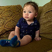 Mateo is registered to the contest to win money with this photo: baby, child, couch, sofa, socks, blue_socks, shirt, navy_shirt, patterned_clothing, indoor, living_room, cushion, brown_couch, seated, curious_expression, infant, person, young_child, skin, hand