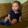 baby, child, couch, sofa, socks, blue_socks, shirt, navy_shirt, patterned_clothing, indoor, living_room, cushion, brown_couch, seated, curious_expression, infant, person, young_child, skin, hand