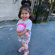 Estella is registered to the contest to win money with this photo: child, toddler, smiling, outdoor, toy, stuffed_animal, concrete, sidewalk, fence, wall, hair_tied, pink_clothing, shoes, happy, person, daylight, playful, cute, holding, casual_wear
