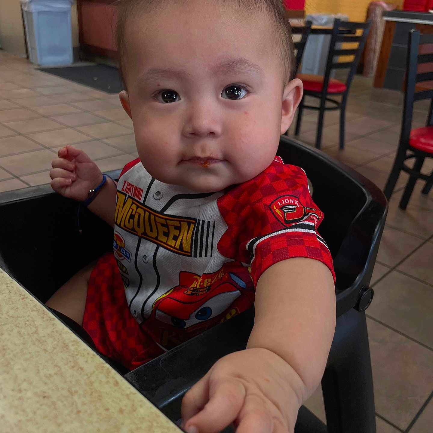 Jose is registered to the contest to win money with this photo: baby, black_chair, bright_colors, casual, child, curious_expression, cute, food_smudge, high_chair, indoor, lightning_mcqueen, person, portrait, reaching_hand, red_clothing, restaurant, seat, table, tile_floor, young_child