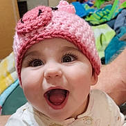 Aela joined the competition — help win amazing prizes! baby, child, infant, smiling, open_mouth, pink_hat, crocheted_hat, big_eyes, long_eyelashes, cute, tongue, teeth, closeup, portrait, happy, indoor, blanket, bedroom, person, arm
