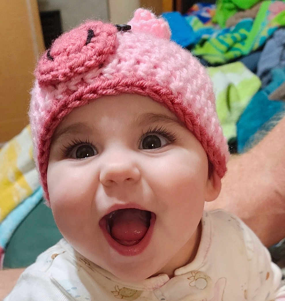 Aela joined the competition — help win amazing prizes! baby, child, infant, smiling, open_mouth, pink_hat, crocheted_hat, big_eyes, long_eyelashes, cute, tongue, teeth, closeup, portrait, happy, indoor, blanket, bedroom, person, arm