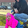 adult_male, affectionate_interaction, baby, black_hoodie, boots, crawling, daylight, grass, jeans, outdoor, parked_car, person, pink_beanie, pink_jacket, sidewalk, smiling, sunglasses, toddler, urban_building, watch
