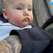 Arlo joined the competition — help win amazing prizes! baby, child, face, blue_eyes, cheeks, towel, tattoo, arm, clothing, black_sleeve, portrait, closeup, skin, expression, person, indoors, soft_light, blurred_background, curly_hair, gaze