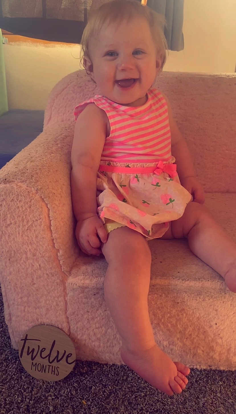 Paige is registered to the contest to win money with this photo: baby, child, smiling, pink_dress, striped_clothing, floral_pattern, chair, soft_furniture, carpet, indoor, twelve_months_sign, happy, cute, one_year_old, infant, sitting, foot, hand, skin, portrait