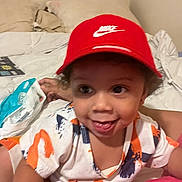 Brandon joined the competition — help win amazing prizes! toddler, child, red_cap, nike, shirt, bed, blanket, smile, tongue_out, indoor, person, happy, face, curly_hair, colorful_clothing, playful, hand, pillow, room, casual