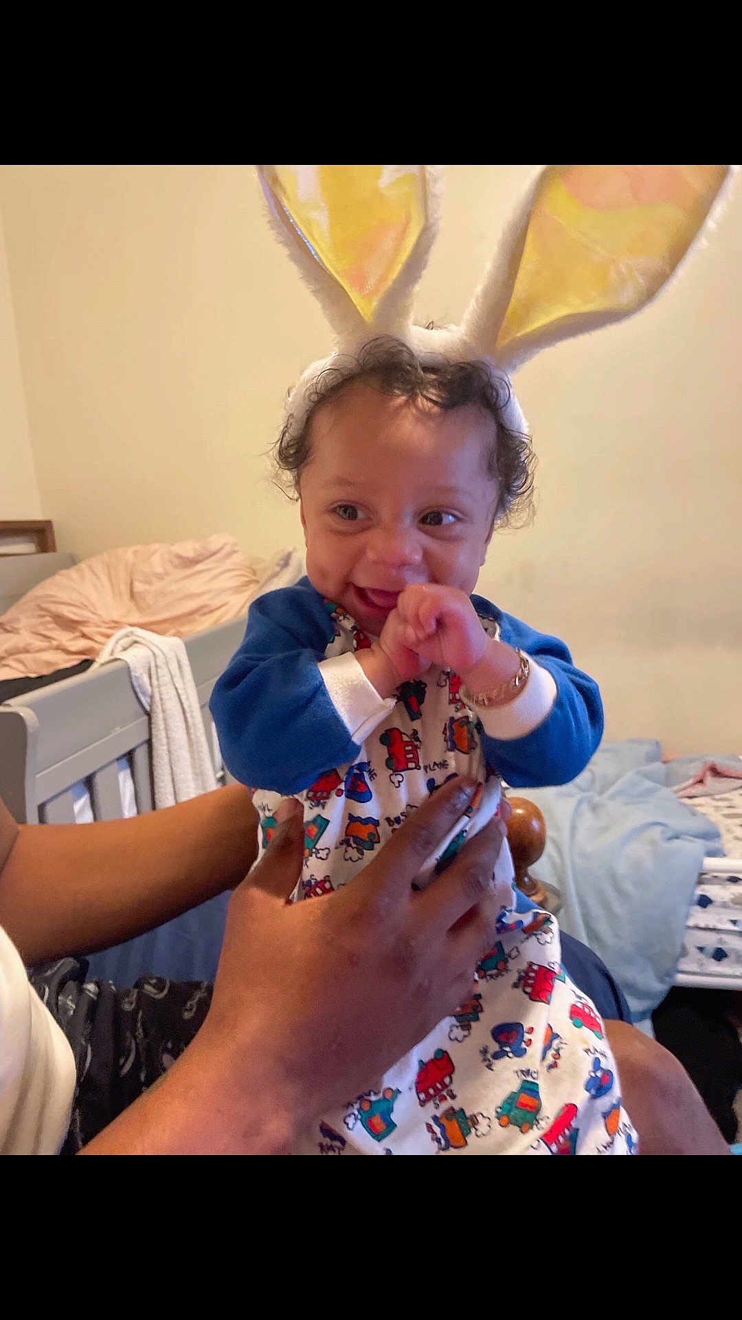 Brandon is registered to the contest to win money with this photo: baby, bunny_ears, smiling, pajamas, child, indoor, bedroom, person, hands, cute, happy, infant, curly_hair, bracelet, furniture, bed, blanket, toy_pattern, playful, holding