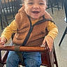 baby, child, smiling, happy, portrait, high_chair, restaurant, cafe, seated, curly_hair, fleece_vest, yellow_shirt, blue_jeans, boots, wooden_chair, furniture, indoor, table, background, photography