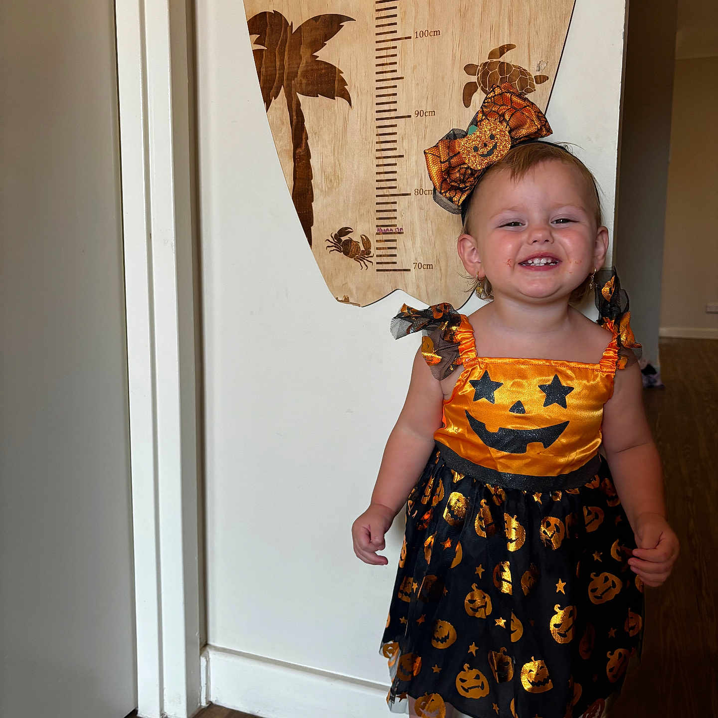 Ahana-Opal is registered to the contest to win money with this photo: carpet, child, decor, door, dress, earrings, floor, footwear, halloween, happy, headband, height_chart, indoors, person, pumpkin, smiling, standing, toddler, wall, wood_floor