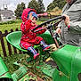 child, toddler, tractor, green, red_rain_suit, boots, steering_wheel, adult, outdoor, farm, grass, fence, trees, overcast_sky, playful, smiling, person, clothing, vehicle, rustic