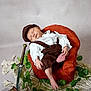 baby, sleeping, brown_clothing, vintage_outfit, cap, orange_chair, toy_sword, treasure_chest, gold_rings, gold_coins, green_leaves, cream_rug, indoor, portrait, peaceful, infant, props, decorative, cute, resting