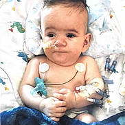 Callan is registered to the contest to win money with this photo: baby, infant, child, hospital, hospital_bed, medical_equipment, monitoring, feeding_tube, nasal_tube, iv, medical_sensors, blanket, pillow, sitting, portrait, face, dark_hair, eyes_open, indoor, healthcare