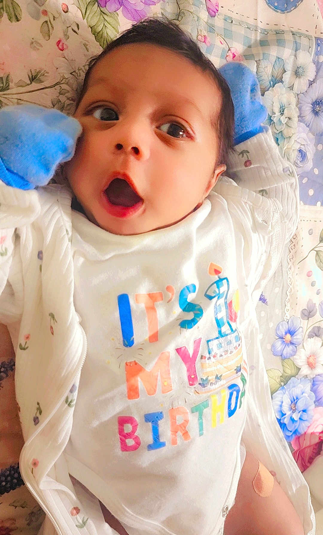 Yousuf joined the competition — help win amazing prizes! baby, infant, mittens, clothing, onesie, birthday, face, expression, lying_down, blanket, floral_pattern, cute, child, person, celebration, happy, closeup, skin, head, mouth_open