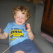 Ace joined the competition — help win amazing prizes! child, toddler, curly_hair, blue_tshirt, school_bus_graphic, shorts, smiling, indoor, armchair, grey_furniture, playing, happy, person, casual_clothing, young, relaxed, home, blurry_hand, seated, cute
