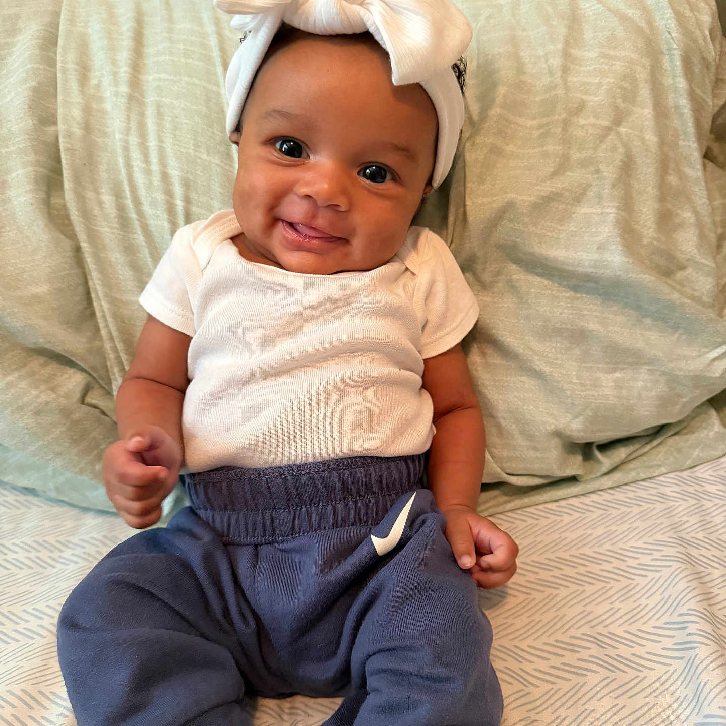 Oak'Lynn is registered to the contest to win money with this photo: baby, child, smiling, headband, bow, white_shirt, blue_pants, sitting, bed, pillow, blanket, happy, indoors, portrait, infant, cute, person, clothing, young_child, face