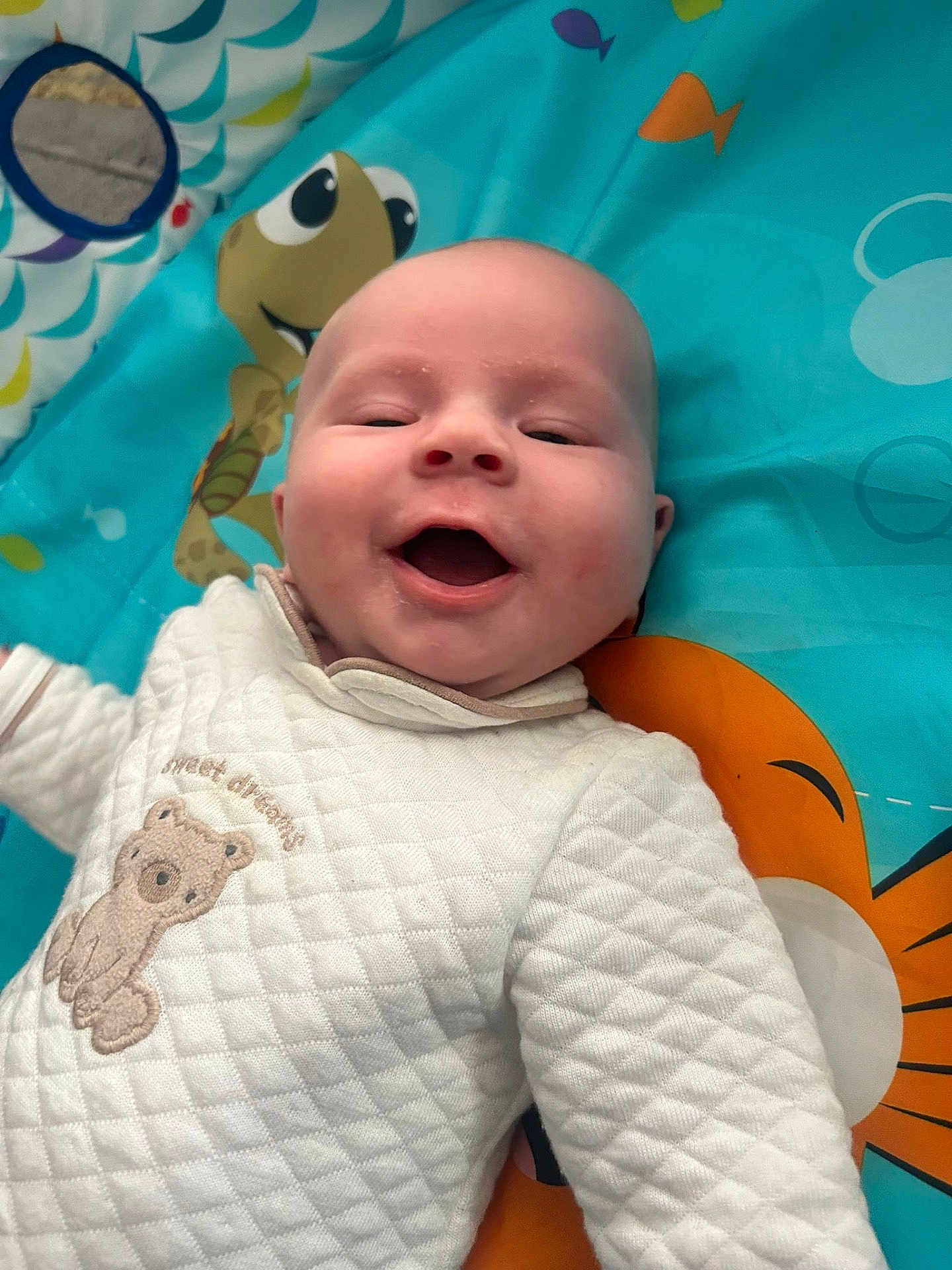 Lillie-Mai is registered to the contest to win money with this photo: baby, smiling, infant, play_mat, quilted_clothing, bear_embroidery, happy, face, child, cute, indoors, colorful, cartoon, turtle, orange, blue, white, person, portrait, joyful