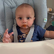 Grey is registered to the contest to win money with this photo: baby, child, infant, high_chair, feeding, sweet_potato, food, orange_smudge, sitting, portrait, indoor, home, blue_shirt, harness, tray, patterned_chair, wide_eyes, facial_expression, first_food, family_moment