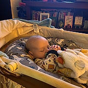 Dominic is registered to the contest to win money with this photo: baby, sleeping, bassinet, blanket, animal_pattern, indoors, cozy, child, infant, pacifier, soft_toys, furniture, television, bookshelf, decor, warm_lighting, rest, peaceful, portrait, home