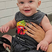Dean is registered to the contest to win money with this photo: baby, child, smiling, sitting, hand, sleeveless_shirt, car_print, saddle, carving, brick_wall, happy, person, cute, infant, outdoor, skin, face, portrait, holding, casual