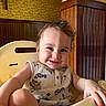 toddler, child, smile, high_chair, sleeveless_shirt, patterned_clothing, indoor, wood_paneling, yellow, cute, happy, portrait, baby, furniture, seat, person, skin, face, hand, hair