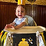 baby, child, crown, high_chair, birthday, party, decorations, balloon, smile, happy, face, clothing, banner, yellow, black, white, indoor, wood_paneling, celebration, toddler