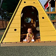 Evelyn is registered to the contest to win money with this photo: baby, child, clothing, daytime, fun, happy, outdoor, park, person, playground, playhouse, playtime, recreation, sitting, smiling, sock, structure, sunlight, wood, yellow