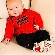 Jude is registered to the contest to win money with this photo: baby, infant, child, couch, red_sweatshirt, text_on_clothing, smile, chubby_cheeks, blue_eyes, sneakers, sitting, portrait, hand, pale_skin, pillow, indoors, fashion, casual, cute, toddler