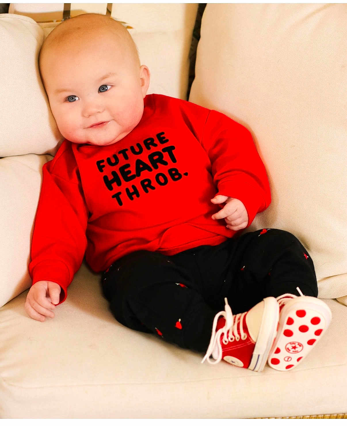 Jude is registered to the contest to win money with this photo: baby, infant, child, couch, red_sweatshirt, text_on_clothing, smile, chubby_cheeks, blue_eyes, sneakers, sitting, portrait, hand, pale_skin, pillow, indoors, fashion, casual, cute, toddler