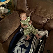 Jersey is registered to the contest to win money with this photo: baby, child, smiling, green_outfit, recliner, blanket, indoors, furniture, cozy, person, happy, face, infant, relaxing, home, cushion, footwear, toy, table, bottle