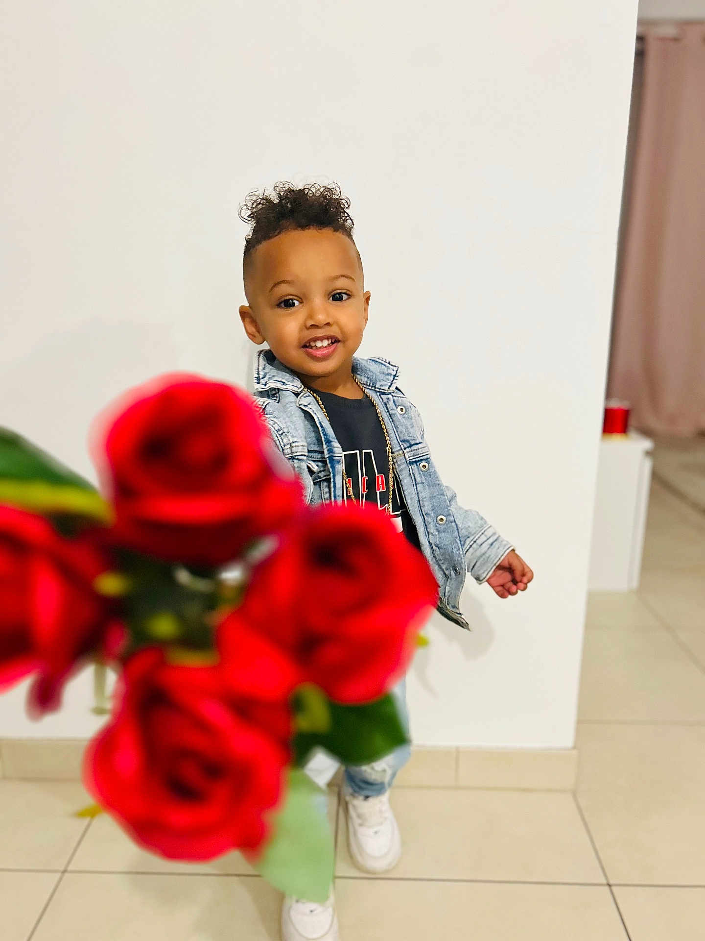 Kendy participe au concours pour gagner de l'argent avec cette photo : blurred_foreground, bouquet, casual_clothing, child, curly_hair, cute, denim_jacket, flowers, happy, holding, indoor, person, portrait, red_roses, smiling, sneakers, tile_floor, toddler, white_wall, young_child
