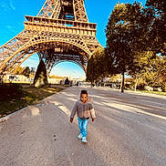 Kendy participe au concours pour gagner de l'argent avec cette photo : architecture, blue_sky, child, daylight, eiffel_tower, happy, jacket, jeans, landmark, nature, outdoor, park, path, shadow, smiling, sneakers, tourist, travel, tree, walking