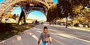 Kendy participe au concours pour gagner de l'argent avec cette photo : architecture, blue_sky, child, daylight, eiffel_tower, happy, jacket, jeans, landmark, nature, outdoor, park, path, shadow, smiling, sneakers, tourist, travel, tree, walking