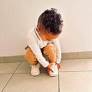 Kendy participe au concours pour gagner de l'argent avec cette photo : child, toddler, curly_hair, white_shoes, sneakers, brown_pants, white_shirt, floor_tiles, indoor, squatting, hands, concentration, young_child, casual_clothing, haircut, candid, cute, person, footwear, minimalist_background