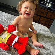 Zion joined the competition — help win amazing prizes! baby, child, curly_hair, diaper, smiling, plush_toy, red_toy, blanket, bedroom, furniture, television, happy, sitting, indoor, cute, infant, person, feet, face, home
