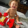 baby, child, curly_hair, diaper, smiling, plush_toy, red_toy, blanket, bedroom, furniture, television, happy, sitting, indoor, cute, infant, person, feet, face, home