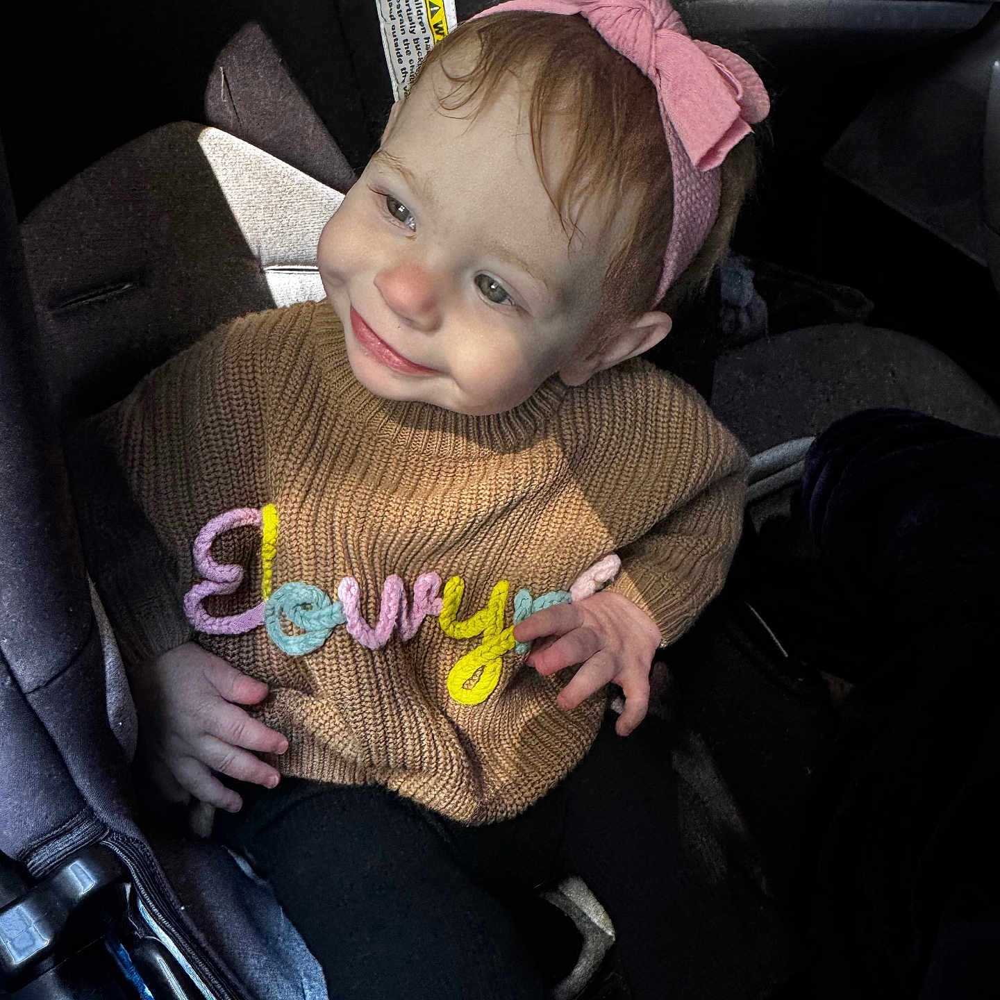 Elowyn is registered to the contest to win money with this photo: baby, brown_sweater, car_seat, child, clothing, colorful_letters, cute, hand, happy, headband, indoor, infant, person, pink_bow, portrait, seatbelt, smiling, sunlight, sweater, toddler
